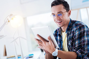 New device. Attractive brunette keeping smile on his face and holding modern gadget in left hand while using it