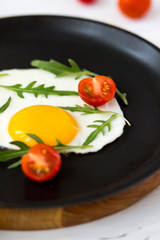 Chicken fried egg in a black frying pan with cherry tomatoes, arugula, salt. Breakfast on a white wooden table. Just cooked food