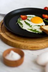 Chicken fried egg in a black frying pan with cherry tomatoes, arugula, salt. Breakfast on a white wooden table. Just cooked food