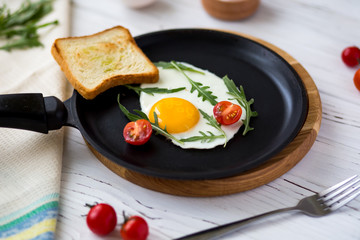 Chicken fried egg in a black frying pan with cherry tomatoes, arugula, salt. Breakfast on a white wooden table. Just cooked food
