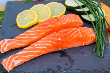 Two pieces of  salmon with onions, lemon, zucchini and green onions on a black board.