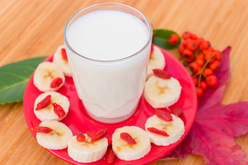 Milk in a glass on a red saucer , chopped banana and goji berries.