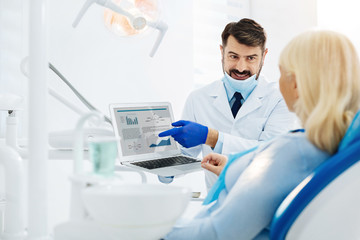 Useful information. Positive experienced dentist holding a laptop in hands while consulting the patient and having positive mood