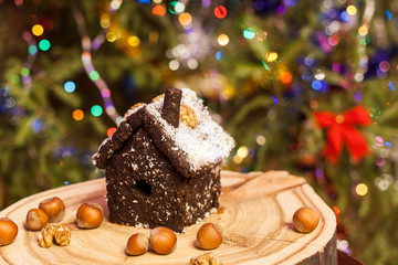 House made of crushed dried prunes, nuts, dates is standing on round wooden board on Christmas tree background.