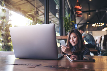 Closeup image of a beautiful Asian business woman holding , using and looking at smart phone with laptop and tablet pc on wooden table in modern cafe