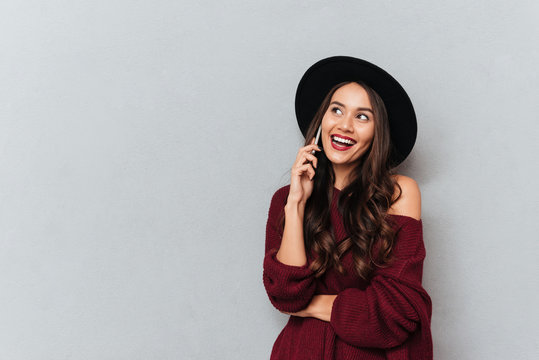 Happy brunette woman in sweater and hat talking by smartphone