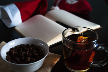 Planning future on Christmas: a notebook with blank pages, a black pen, Santa hat, glass mug with teabag, white bowl of chocolate cereal rings and milk on plain cloth, all on dark dining table