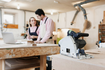 young happy family standing at a work bench in a carpentry workshop, writing a project. Electric plane in the foreground in focus. Family business. startup business. young specialist designer.