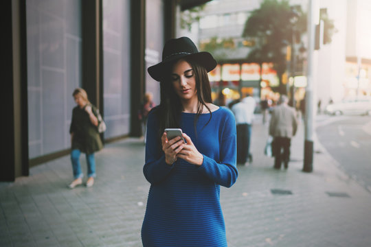Successful freelancer working by a mobile phone connected to wifi while walking the street. Charming hipster girl wearing hat and dress searching the needed address in the internet on a smartphone.