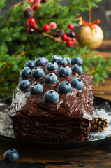 Christmas chocolate cake with fresh blueberries on a black plate on the table decorated with green fir-tree brunches.