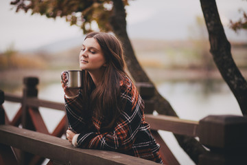 Young beautiful woman in a warm classic coat drinking tea near lake. Autumn atmosphere