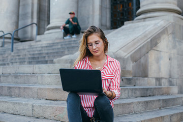 Portrait of casual, fashionable and trendy dressed young millennial woman typing on keyboard or working remotely outdoors, sitting on steps of office building or university, concept young