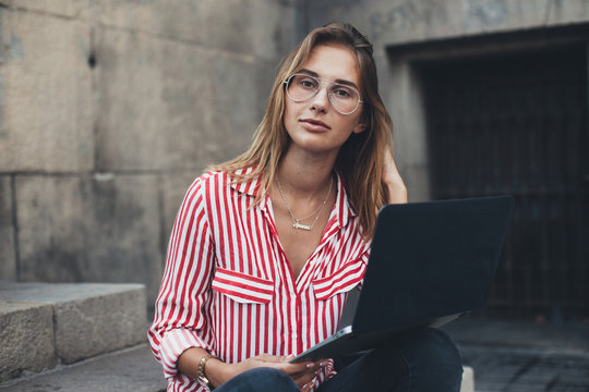 Portrait of casual, fashionable and trendy dressed young millennial woman typing on keyboard or working remotely outdoors, sitting on steps of office building or university, concept young