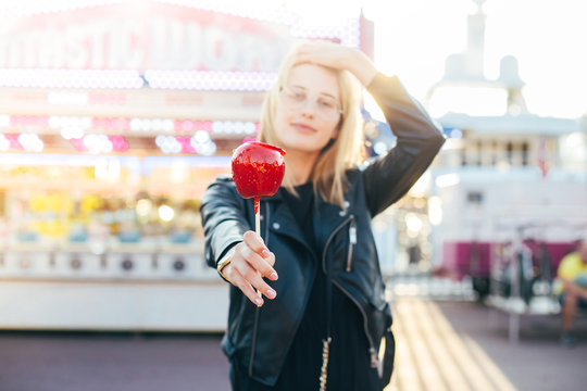 Beautiful young fashion model or lifestyle hipster blogger poses with dreamy and sexy look on her face, sunshine flare in hair, holds holiday candy caramel red apple at amusement park or fair