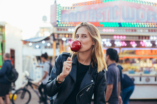 Cute, young female in cool black leather jacket and prescription glasses smiling kissing face expression, holds big red candy toffee apple, stands in middle of crowd in amusement carnival park or fair