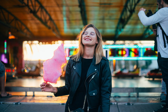 Happy and smiling young woman in cool trendy outfit, hipster or fashion model, enjoys vacations or holidays at amusement park or carnival fair, holds up in sunset light huge pink cotton candy