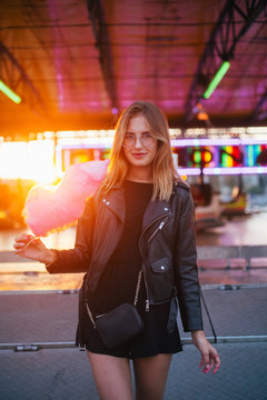 Happy and smiling young woman in cool trendy outfit, hipster or fashion model, enjoys vacations or holidays at amusement park or carnival fair, holds up in sunset pink cotton candy and looks in camera