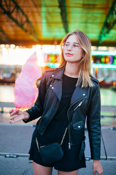 Cute and pretty girl or student, eats and poses with sugar pink candy cotton, wears leather jacket in line for attraction ride or rollercoaster on town festival or fair, concept happy times in summer
