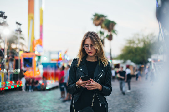 Trendy fashion teenager or young woman uses communication technology or messaging application to keep in touch or meetup with friends on smartphone, in middle of crowd at amusement park 