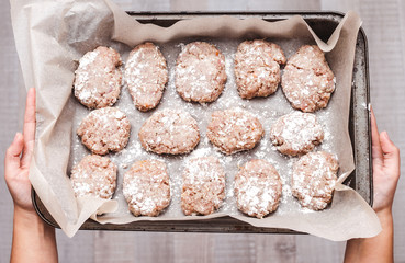 Young housewife holds a tray with fresh minced raw meat cutlets covered with flakes of flour on light wood background, view from above.