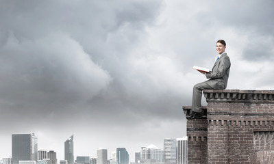Man on roof edge reading book and cityscape at background