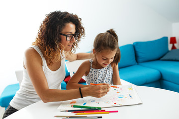 Mother teach her daughter to draw. They're sitting at the table in the living room.