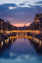 Purple cloudy sky, reflections in the water, boats and cosy-lighted streets during a twilight blue hour sunset, Amsterdam, Netherlands