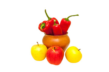 Red peppers in a brown ceramic pot and apples lying around, isolated on white background