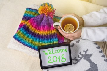 Welcome 2018, text in frame. Winter clothing background and a cup of hot tea with lemon.