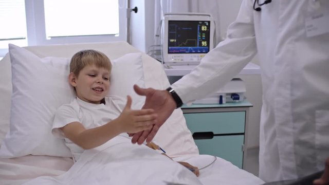 Cute little boy lying on hospital bed, listening to doctor examining x-ray image and shaking hands with him