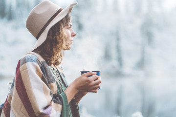 Fototapeta premium Pretty woman with hot drink in hands spending vacations among stunning winter landscape. Traveling in mountains wilderness. Wanderlust and boho style