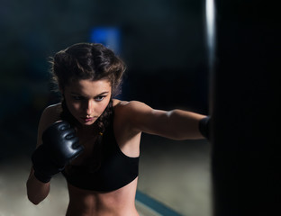 Fototapeta na wymiar Beautiful young fighter boxer fit girl wearing boxing gloves in training, focused on heavy punching bag in gym. She is in good shape. Woman power