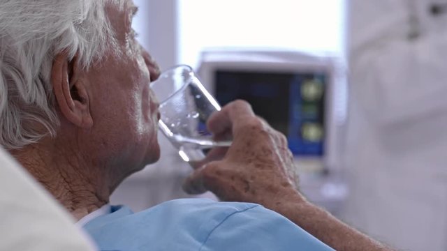 Senior man lying on bed in hospital ward, taking medications and drinking water from glass
