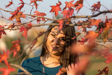 Young beautiful teenage girl smiling behind autumn red leaves on the tree