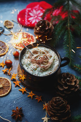 Christmas sparkler in a cup of hot chocolate or cocoa with marshmallows, dry oranges, cinnamon, fur tree, mittens on dark stone background