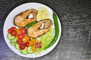 fresh fish, cut into steaks, fried in butter and seasoned with spices, served on a festive white plate with tomatoes, cucumbers, chilli peppers and leeks on a black table as background
