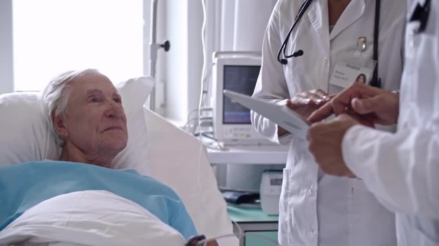 Tilt down of doctors using digital tablet while visiting senior man in hospital ward