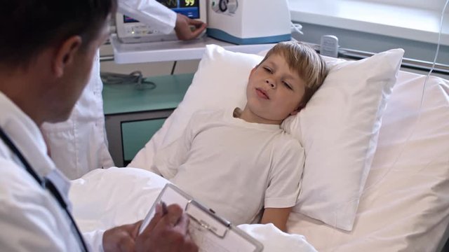 Little blonde boy lying on hospital bed and talking to doctor while he writing down medical card on clipboard