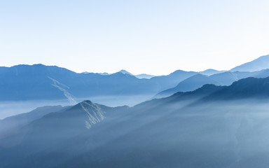 Hazy blue mountains of Zhushan inside Alishan Recreation Area in Taiwan covered by fog during sunrise in morning with bright winter sky.