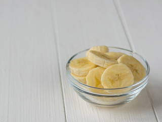 Glass bowl with slices of banana on white wooden table top view.