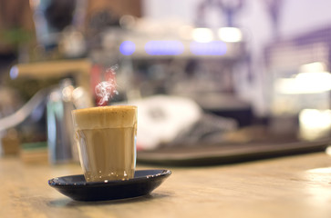 Glass of steaming hot coffee  on a wooden table with blurred bokeh coffee machine background.