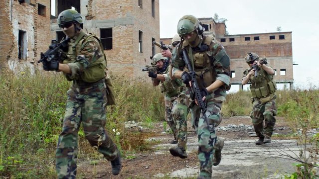 Military squad in camouflage armed with shotguns running together through destroyed area in slow motion
