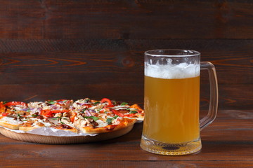 Mug of light beer and pizza with smoked meat and vegetables on dark wooden background.