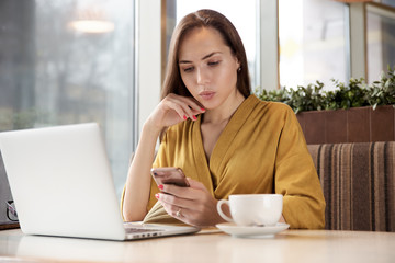 portrait in natural light, the stylish long-haired attractive young woman with smartphone in hand mingling at a table in the cafe with a laptop