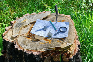 hunting folding knife with steel sharp blade stuck on old tree stump in the wild forest and vintage military topographic map of the area under pencil, stone, compass, magnifying glass