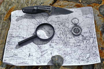 vintage military topographic map of the area on old tree stump under hunting folding knife with steel sharp blade, compass, magnifying glass and stone
