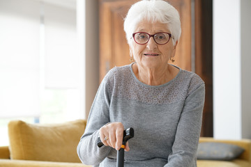 Portrait of elderly woman sitting in sofa, holding cane - obrazy, fototapety, plakaty