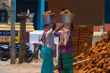 Two Indian women carry heavy bricks clay on their heads in traditional clothes. View from the back. Using female labor at the building. India, Tamil Nadu.
