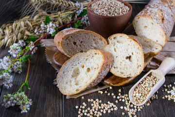 Bakery concept. Buckwheat bread, French baguette and stalks of wheat, oats, buckwheat on dark wooden table.