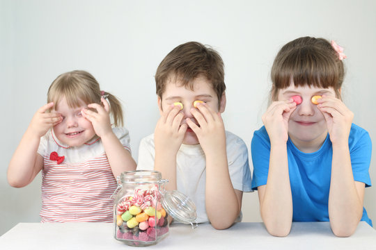 Three happy children close eyes by candies near jar on table in white studio